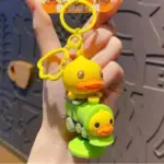 Llavero de silicona con pato amarillo sobre un tren verde y otro patito frontal, con aro y dije de flor amarillos.