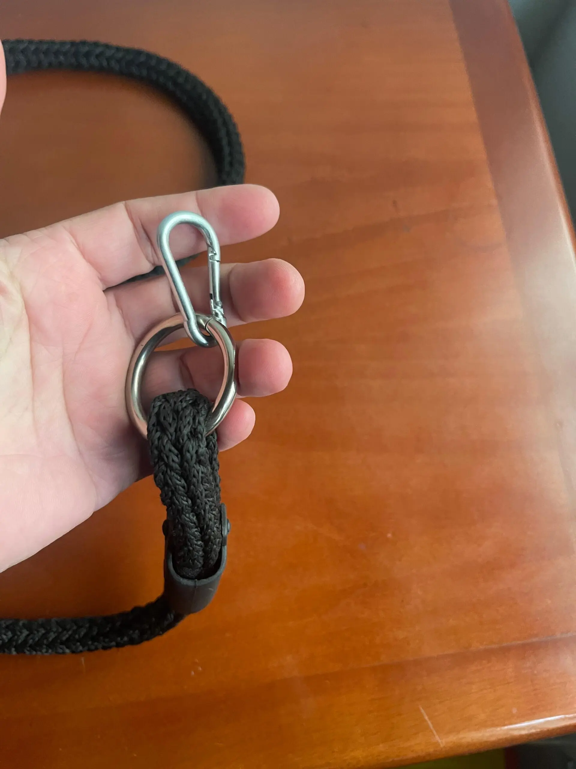 Detalle de correa negra de cuerda trenzada con mosquetón y anillo de metal plateado, sostenida por una mano sobre superficie de madera.