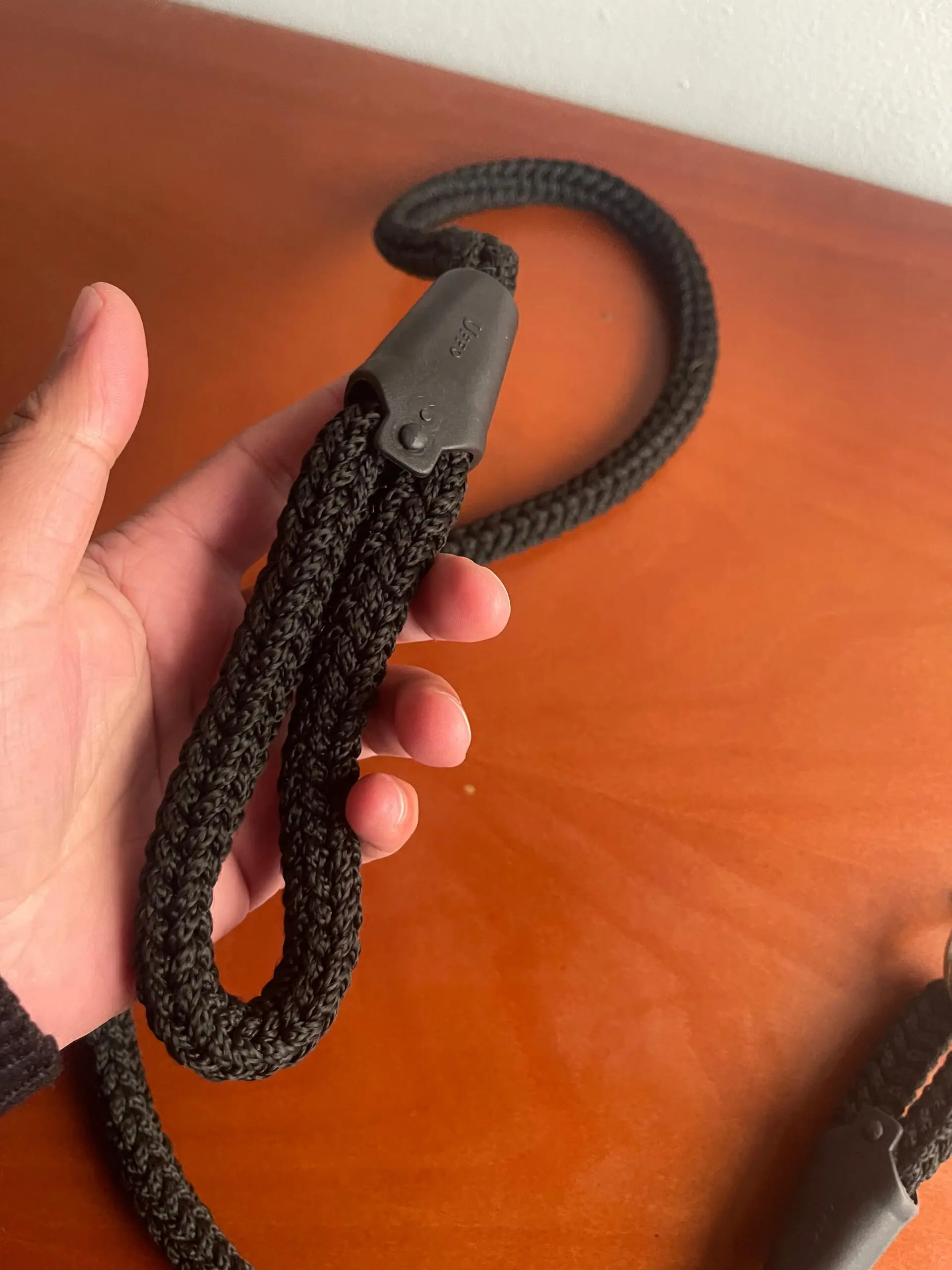 Correa negra de textura trenzada con mango ergonómico de material suave, sostenida por una mano sobre una superficie de madera.