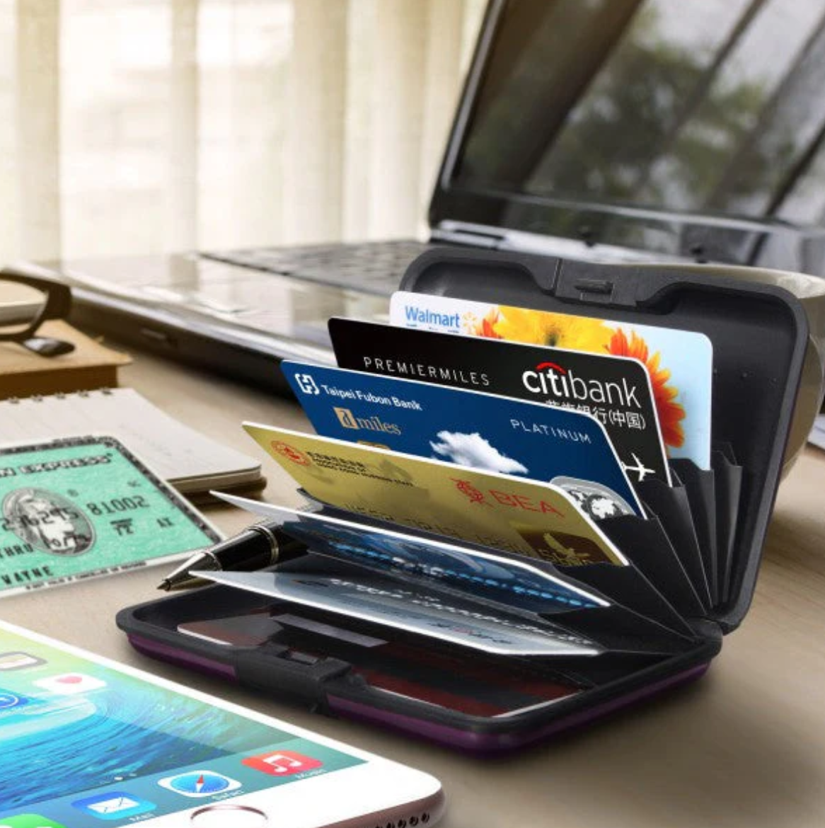 Cartera rígida negra con tarjetas de crédito desplegadas en abanico sobre un escritorio junto a un celular y una laptop.