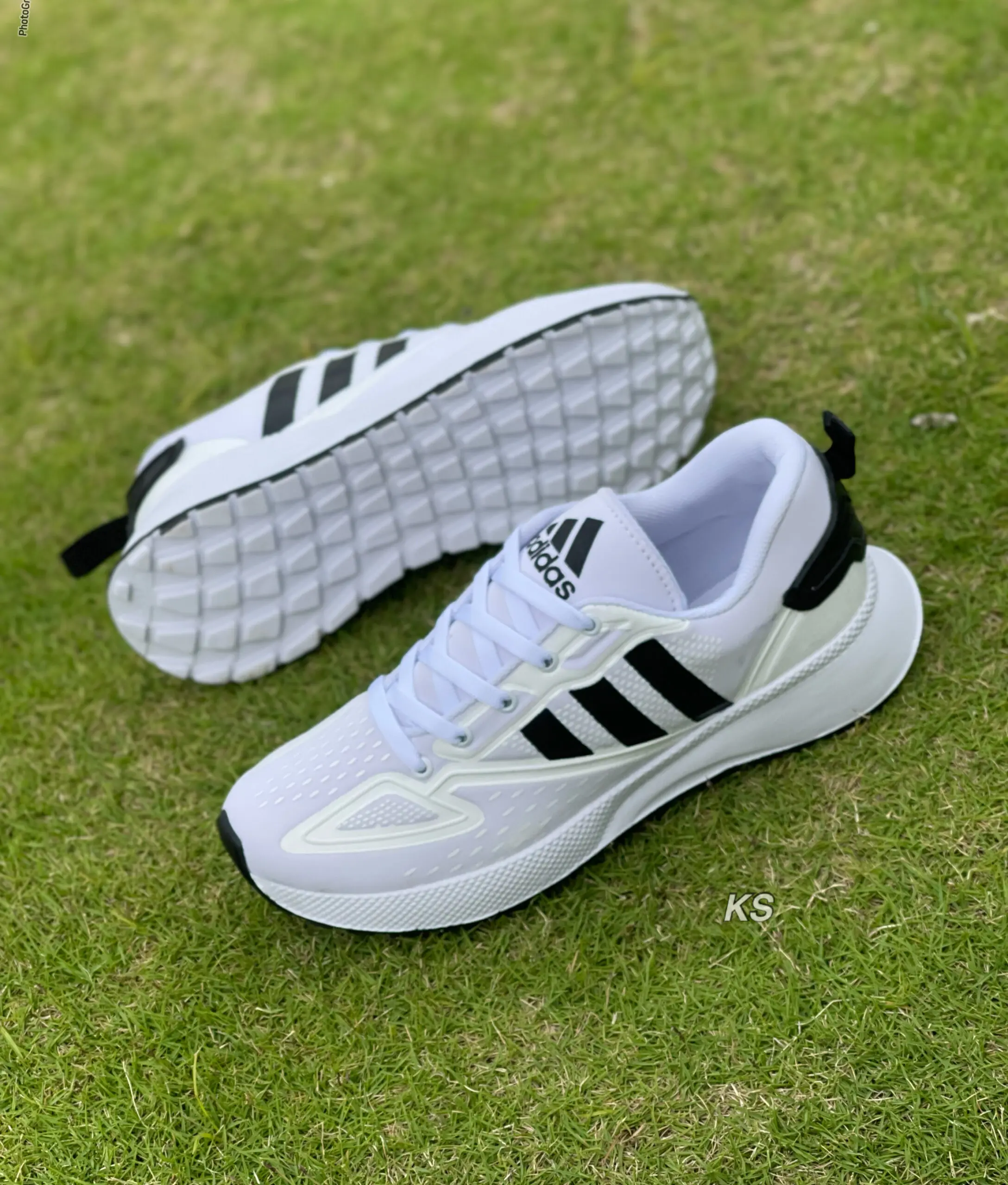 Zapatos deportivos casuales blancos con tres franjas negras, suela flexible texturizada y cordones, ubicados sobre césped verde.