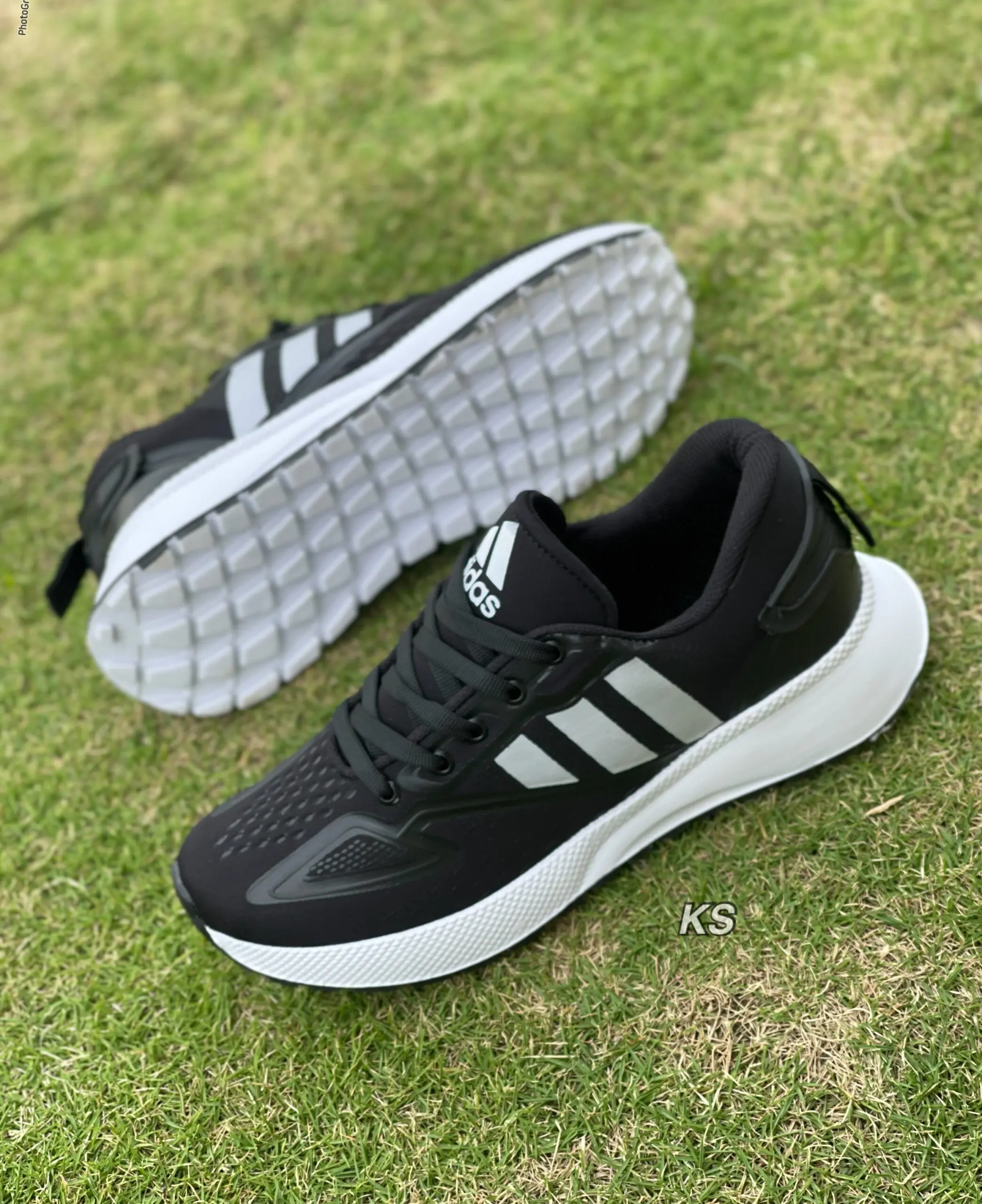 Par de zapatos deportivos negros para hombre con tres franjas plateadas, suela blanca gruesa y flexible, sobre césped verde.