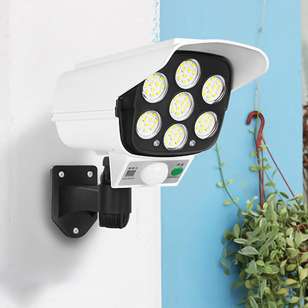 Lámpara solar blanca con diseño de cámara de seguridad, luz LED frontal múltiple, sensor de movimiento y soporte negro montado en pared.