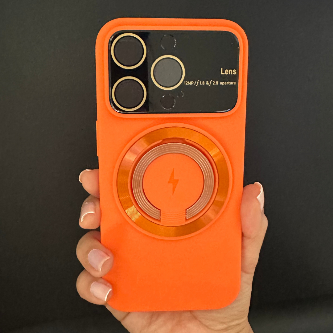 Funda MagSafe de silicona naranja con anillo de carga central y protector de lentes con detalles dorados, sostenida por una mano.