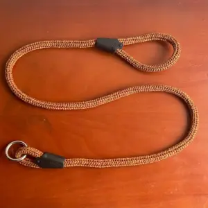 Correa de cuerda trenzada con patrón naranja y amarillo, con mango en lazo, anillo metálico y terminales negros, sobre fondo café.