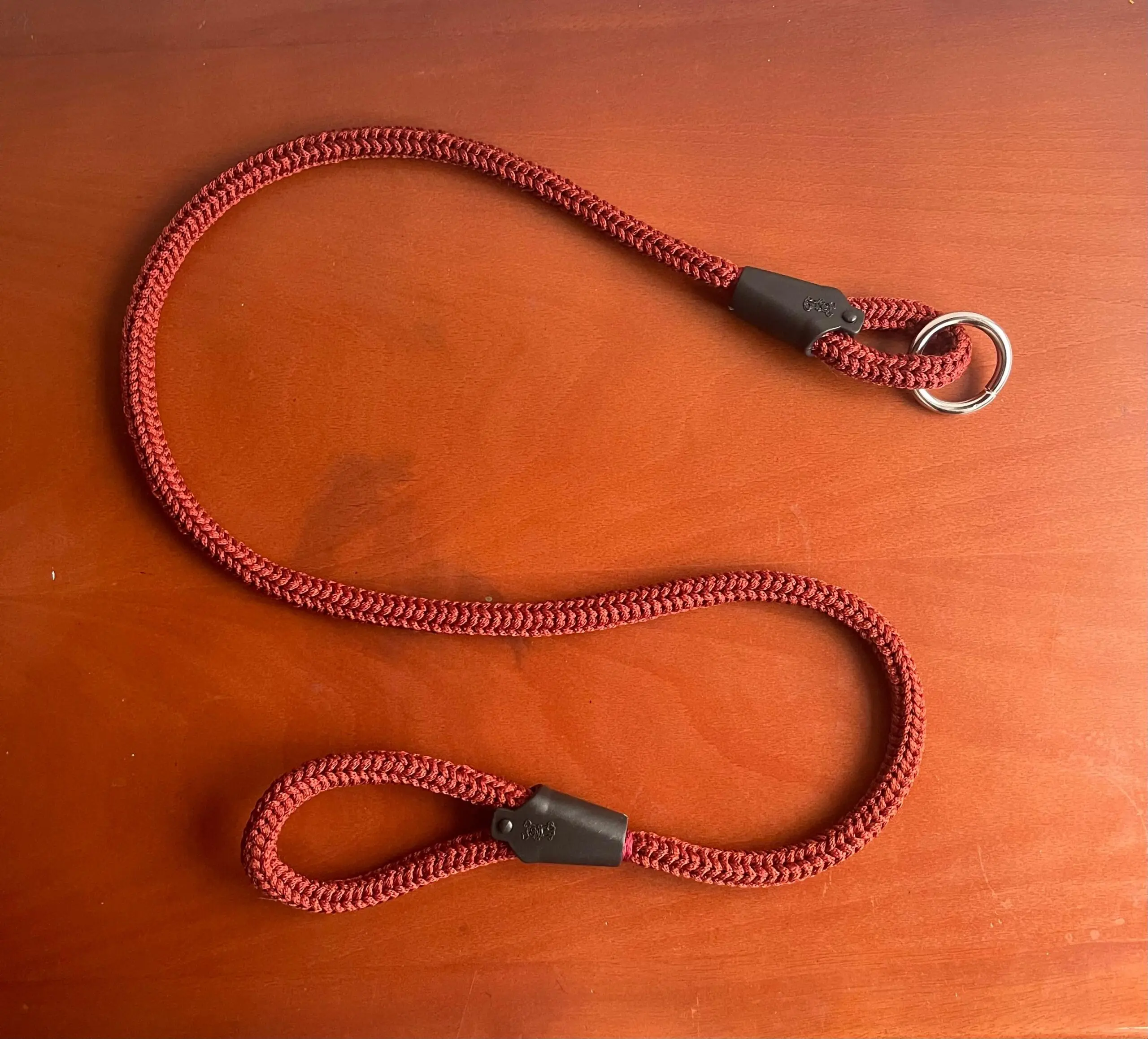 Correa de cuerda trenzada roja con anillo metálico y mango ergonómico con terminales negros, sobre superficie de madera.