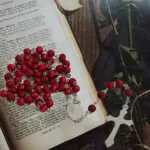 Rosario de cuentas rojas en madera con crucifijo y medalla de acero, colocado sobre un libro abierto junto a una flor.