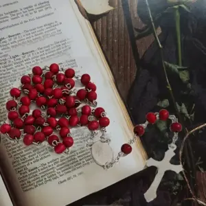 Rosario de cuentas rojas en madera con crucifijo y medalla de acero, colocado sobre un libro abierto junto a una flor.