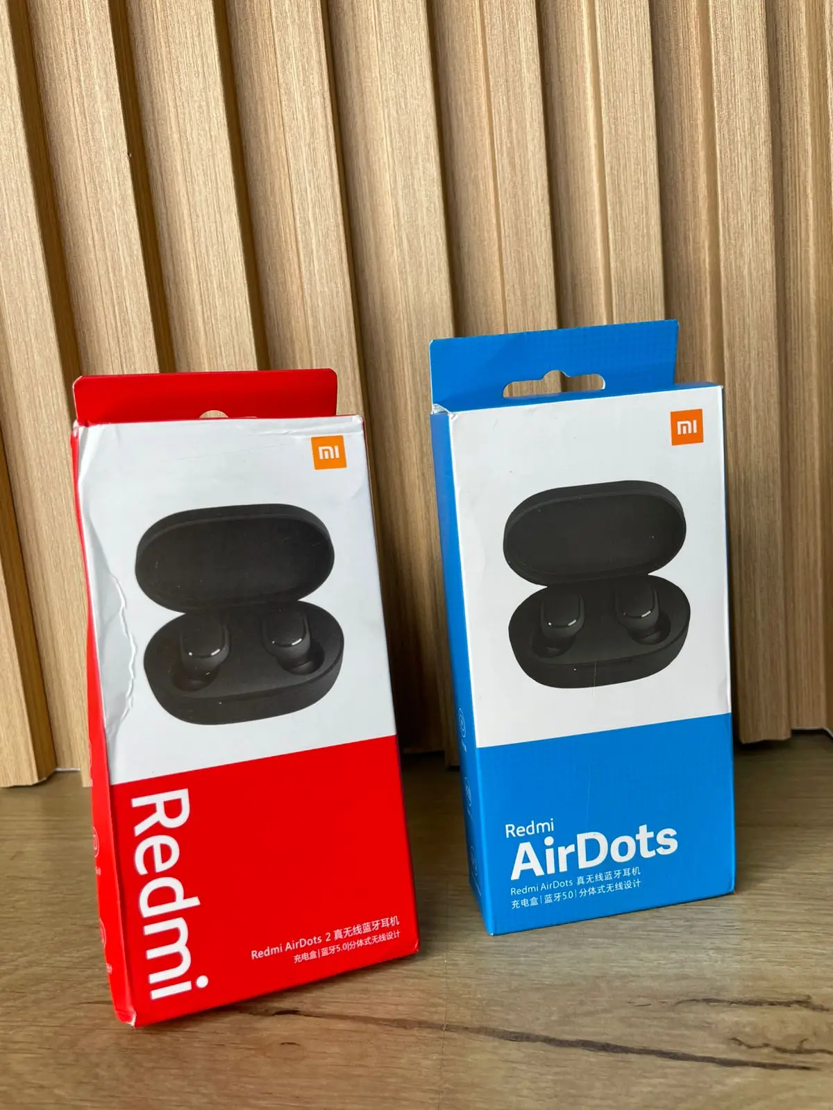 Dos cajas de auriculares Xiaomi Redmi AirDots, una roja y otra azul, sobre una superficie de madera con fondo de listones verticales.
