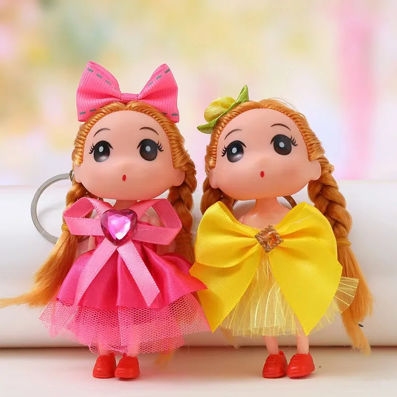 Dos llaveros de muñecas con trenzas, una con vestido rosa y corazón brillante, otra con vestido amarillo y lazo, ideales para decorar bolsos.