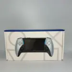 Consola portátil Play X en color plata y negro con detalles azules, frente a su caja blanca con patrones geométricos grises.