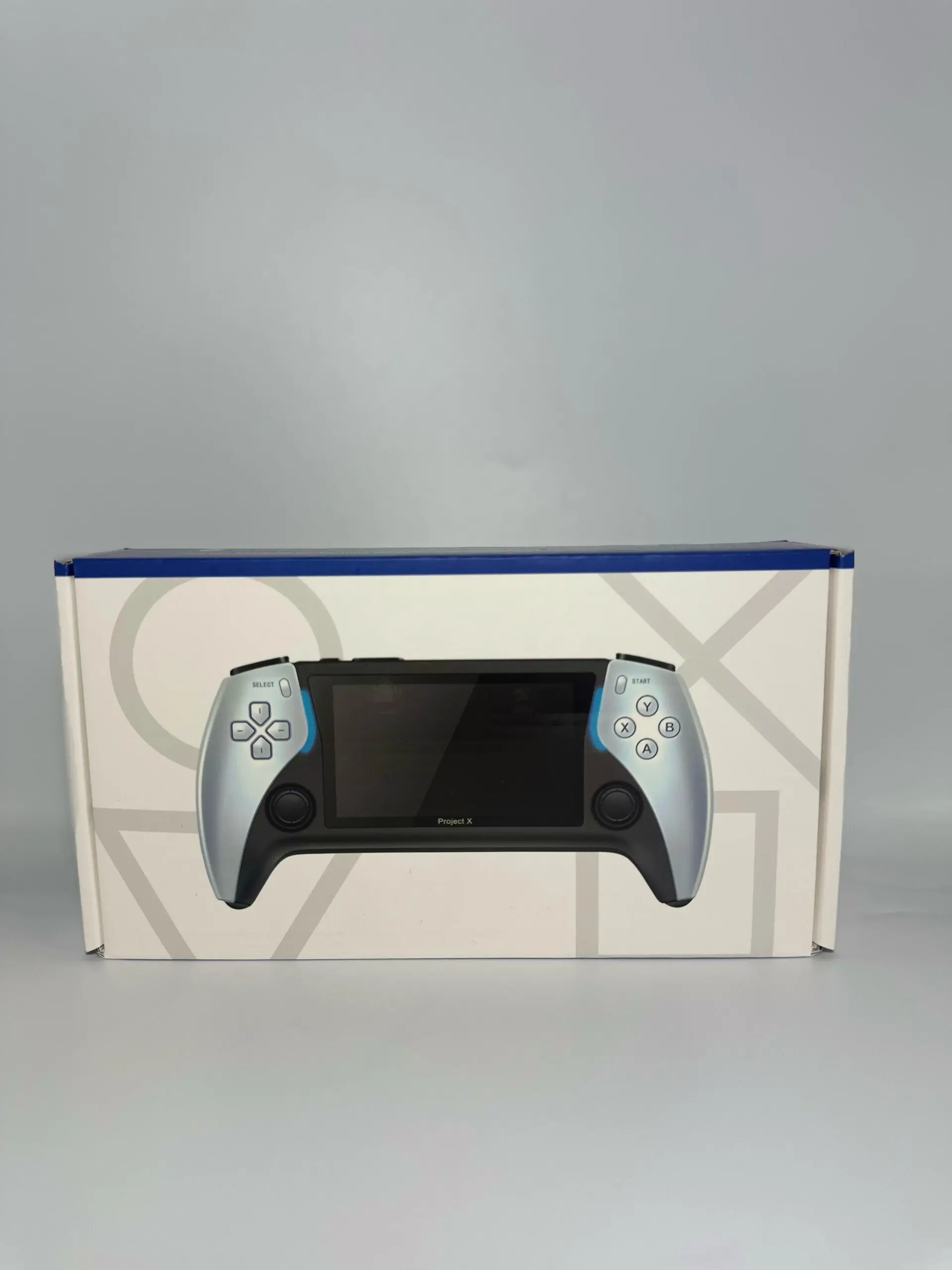 Consola portátil Play X en color plata y negro con detalles azules, frente a su caja blanca con patrones geométricos grises.