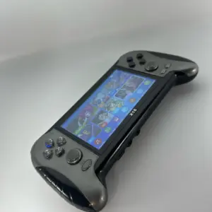 Consola portátil de videojuegos color gris con empuñaduras ergonómicas negras, pantalla táctil encendida y controles físicos en ángulo.