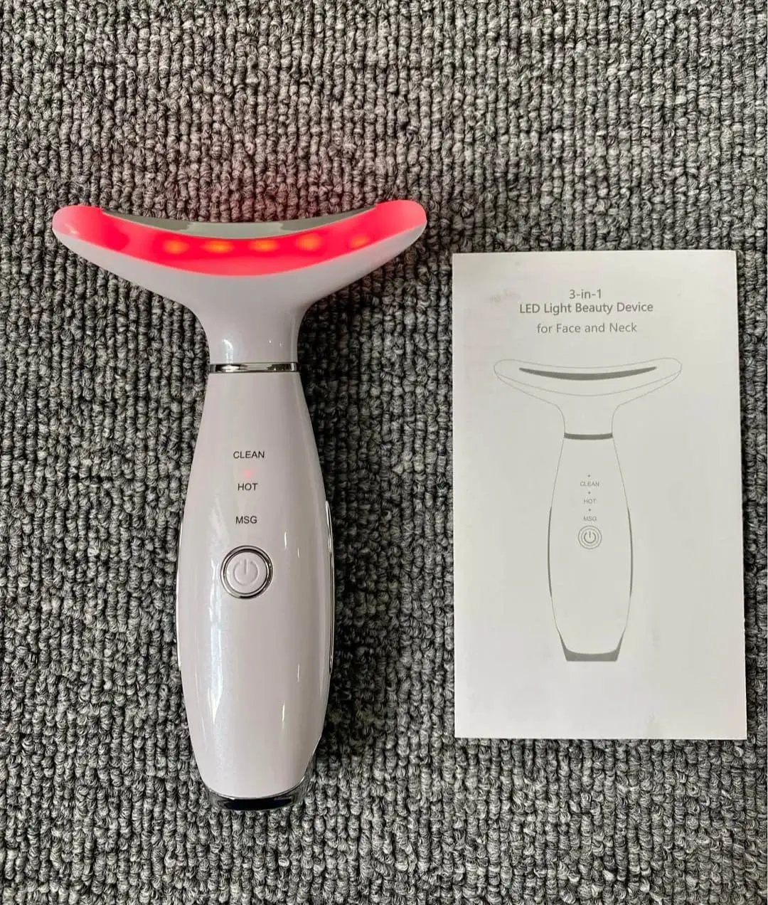 Dispositivo facial y cervical blanco con luz roja encendida, tres botones de control, manual de instrucciones y fondo de textura gris.