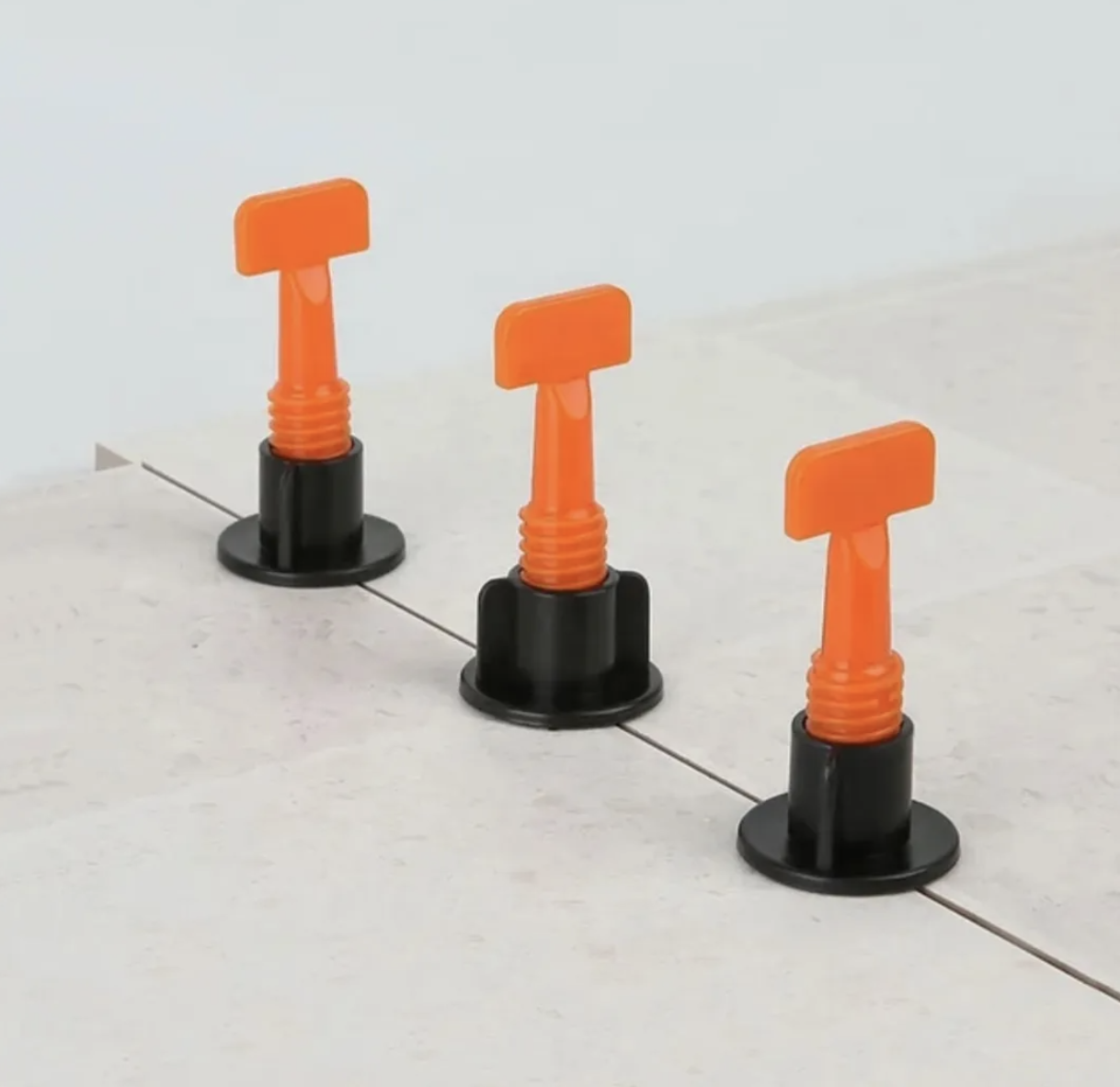 Tres niveladores de cerámica con base negra y vástago roscado naranja, instalados sobre baldosas claras para asegurar una superficie plana y nivelada.