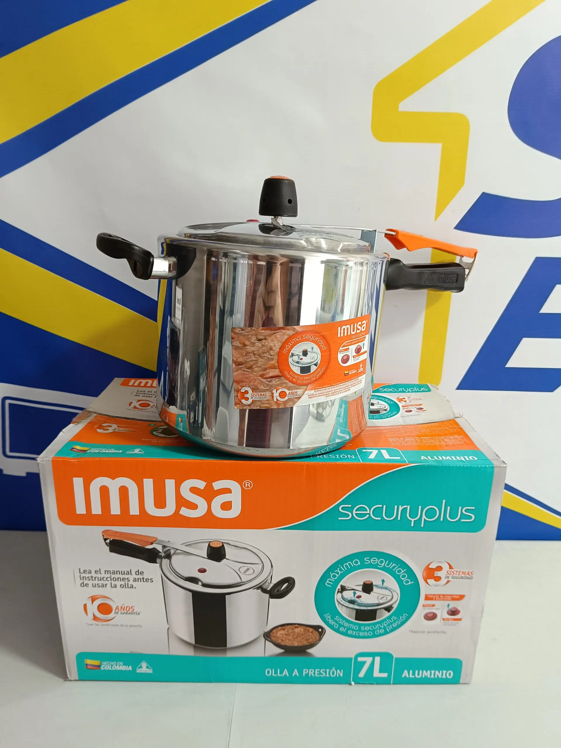 Olla a presión Imusa Secury Plus de 7 litros en aluminio plateado con mangos negros y naranja, sobre su caja de empaque.