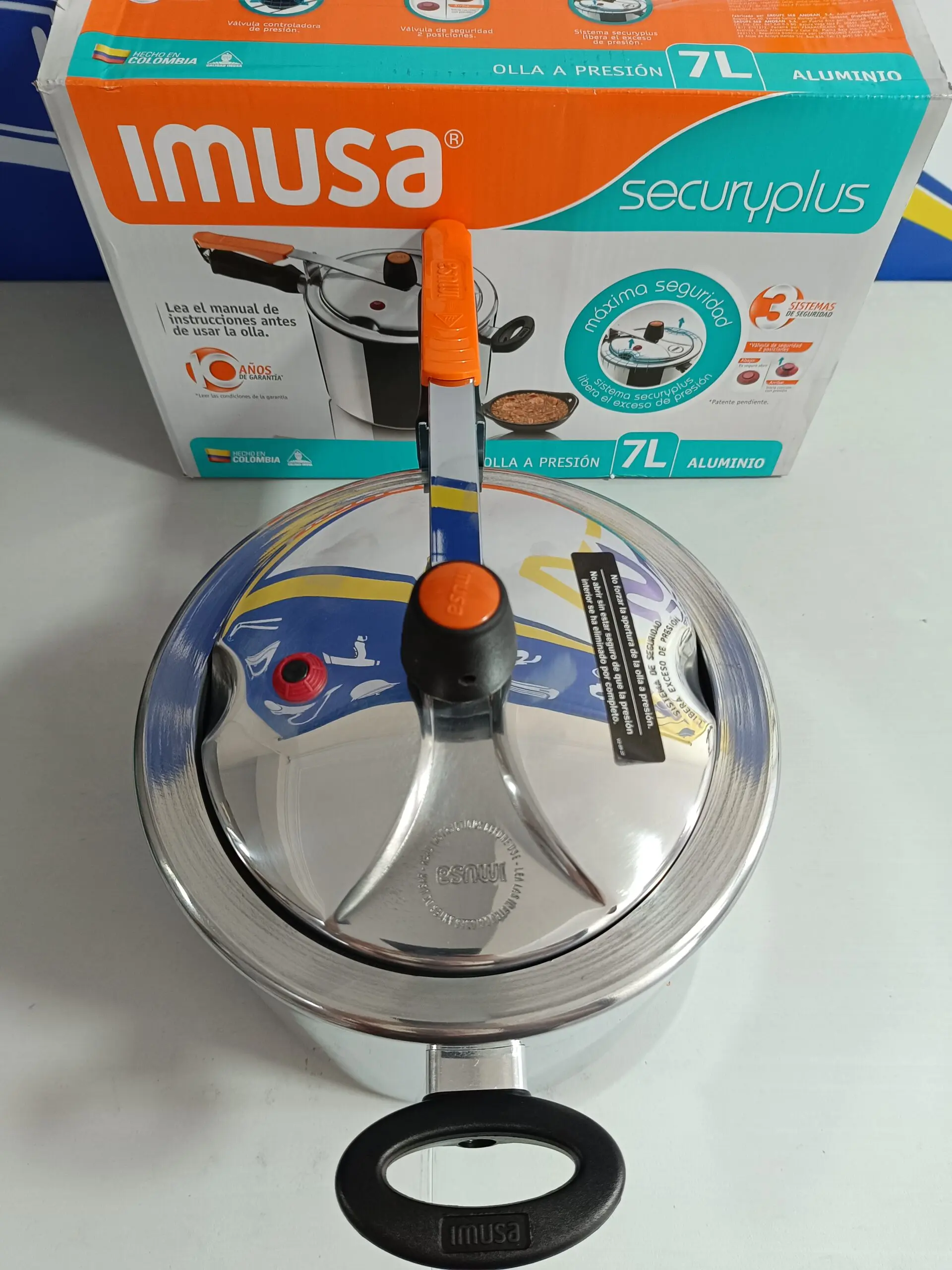 Olla a presión Imusa Secury Plus de 7L en aluminio plateado con tapa de mango naranja, junto a su caja de empaque original.