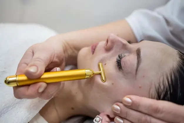 Masajeador facial vibratorio dorado en forma de T siendo aplicado en la mejilla de una mujer para cuidado de la piel.