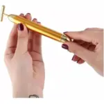 Manos sosteniendo un masajeador facial vibratorio portátil dorado en forma de T con cabezal cilíndrico en el extremo inferior