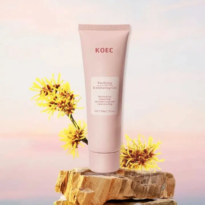 Tubo rosa mate de gel exfoliante purificante de 50g sobre base rocosa, flanqueado por flores amarillas de hamamelis bajo fondo atardecer.