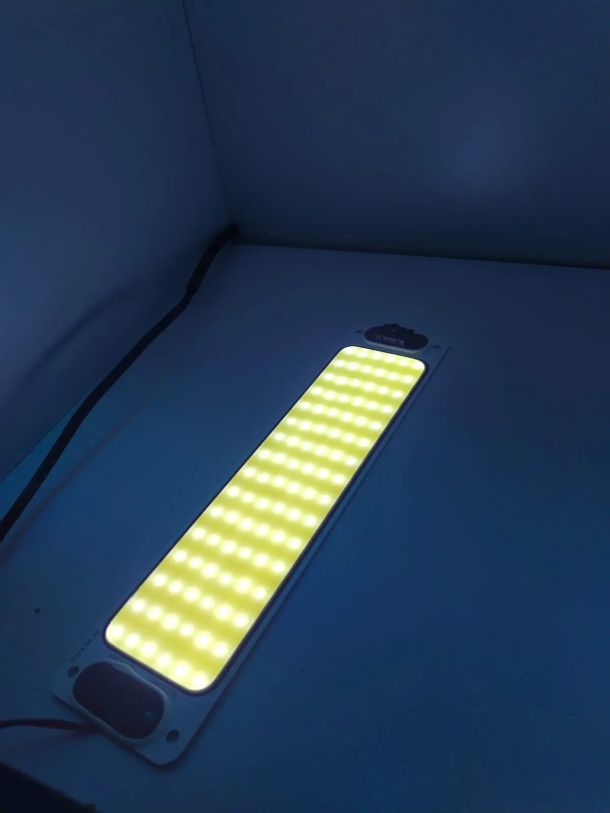Plaqueta LED rectangular para habitáculo de vehículo, encendida con luz brillante sobre superficie oscura, vista en ángulo lateral.