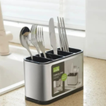 Organizador de cubiertos de acero inoxidable con cuatro compartimentos negros y base, colocado sobre la encimera junto al fregadero, contiene cuchara, tenedores, cuchillo y palillos.