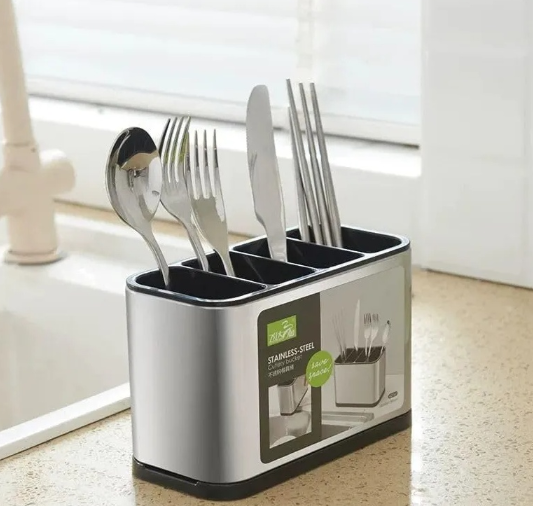 Organizador de cubiertos de acero inoxidable con cuatro compartimentos negros y base, colocado sobre la encimera junto al fregadero, contiene cuchara, tenedores, cuchillo y palillos.