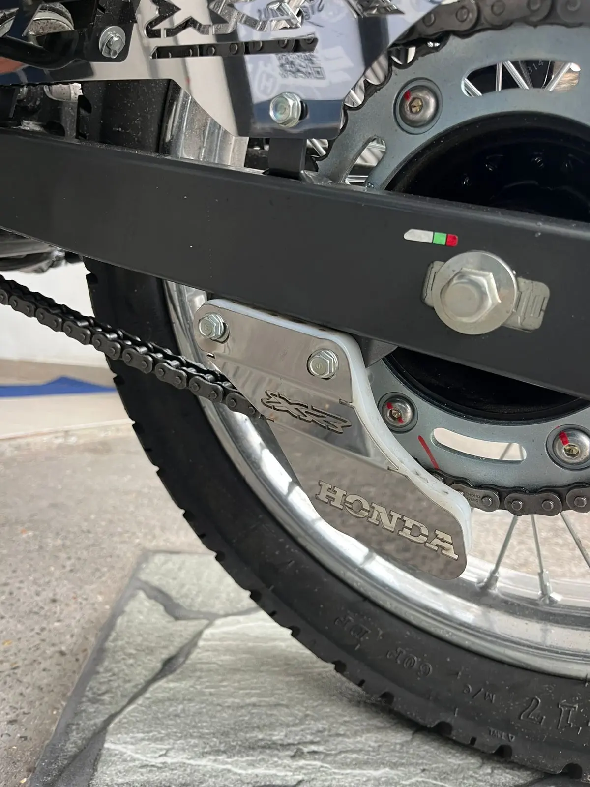 Patín tensor de cadena de acero inoxidable con grabado Honda, instalado en el lateral de la motocicleta para proteger la transmisión trasera.