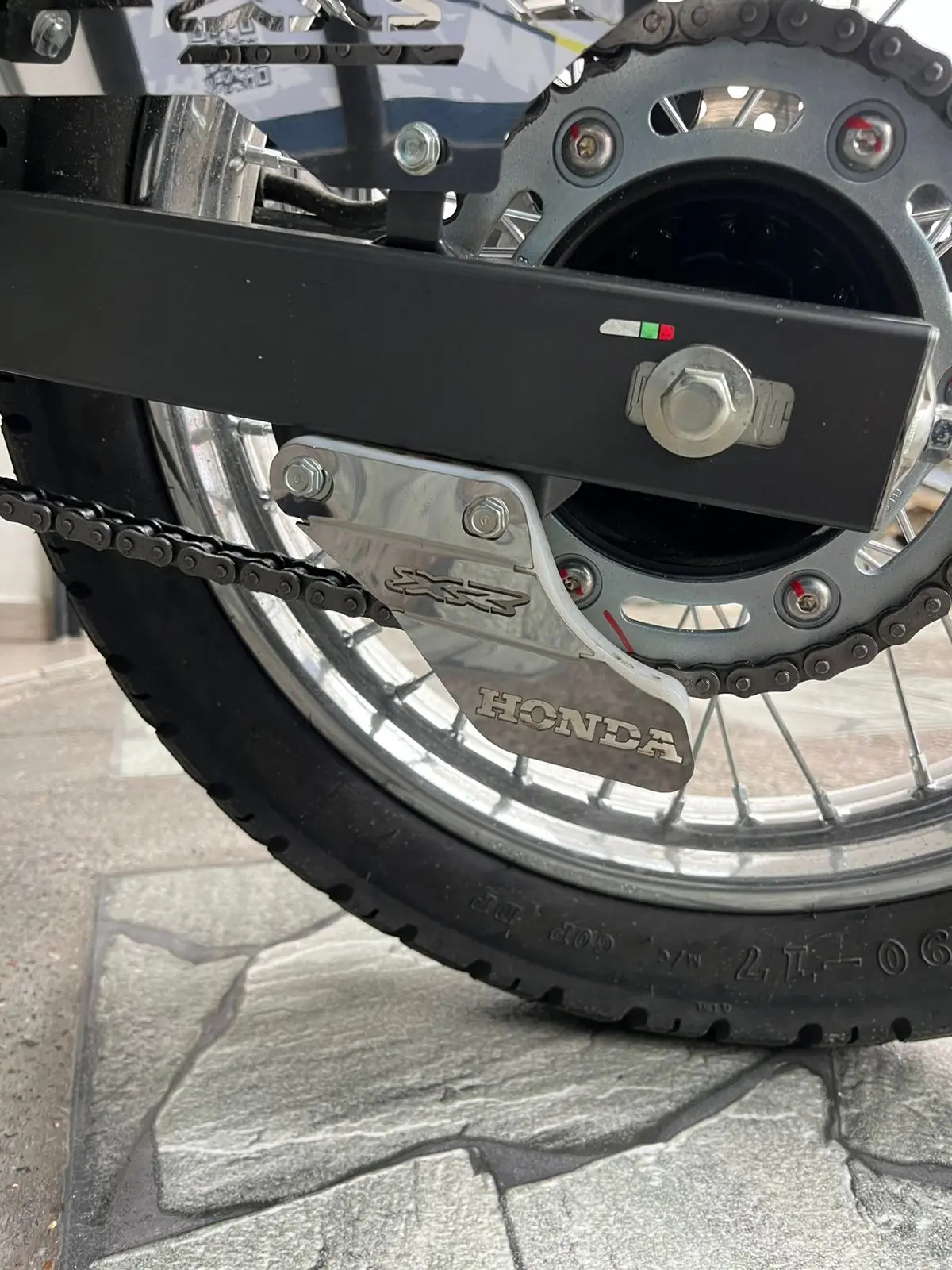 Patín tensor de cadena lateral de acero inoxidable pulido con grabado Honda, instalado en la transmisión trasera de una motocicleta.