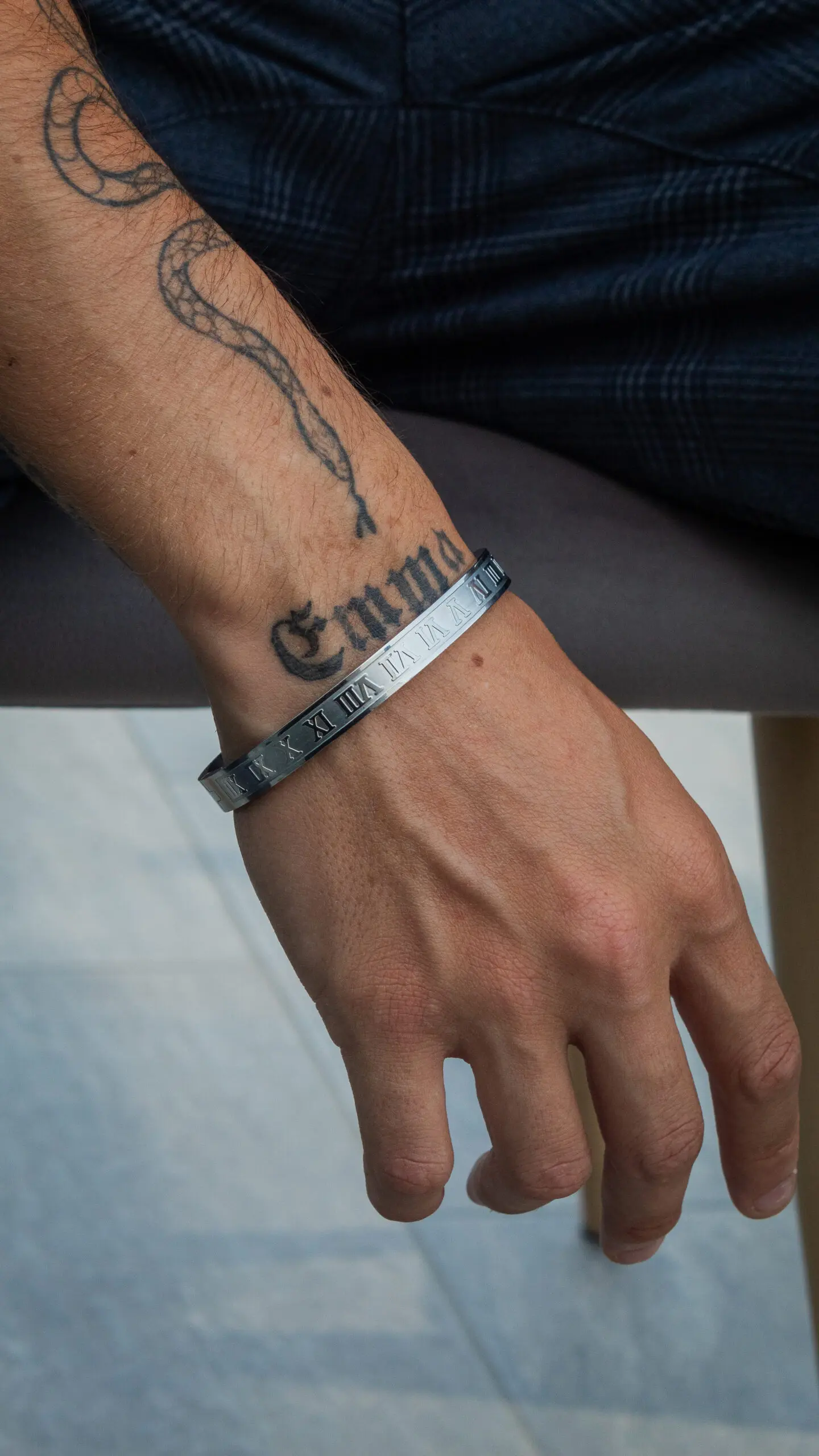 Brazalete rígido de acero plateado con diseño romano grabado, lucido en la muñeca de una persona con tatuajes, resaltando su acabado metálico.