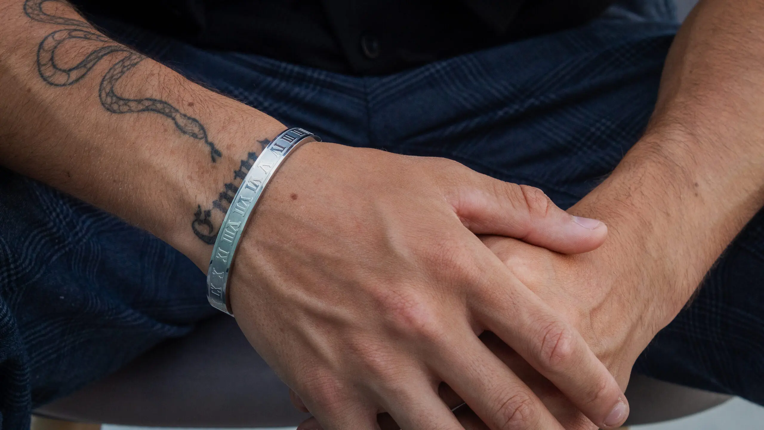 Brazalete rígido de acero plateado con grabados romanos en la muñeca de un hombre, destacando su acabado metálico y diseño elegante.