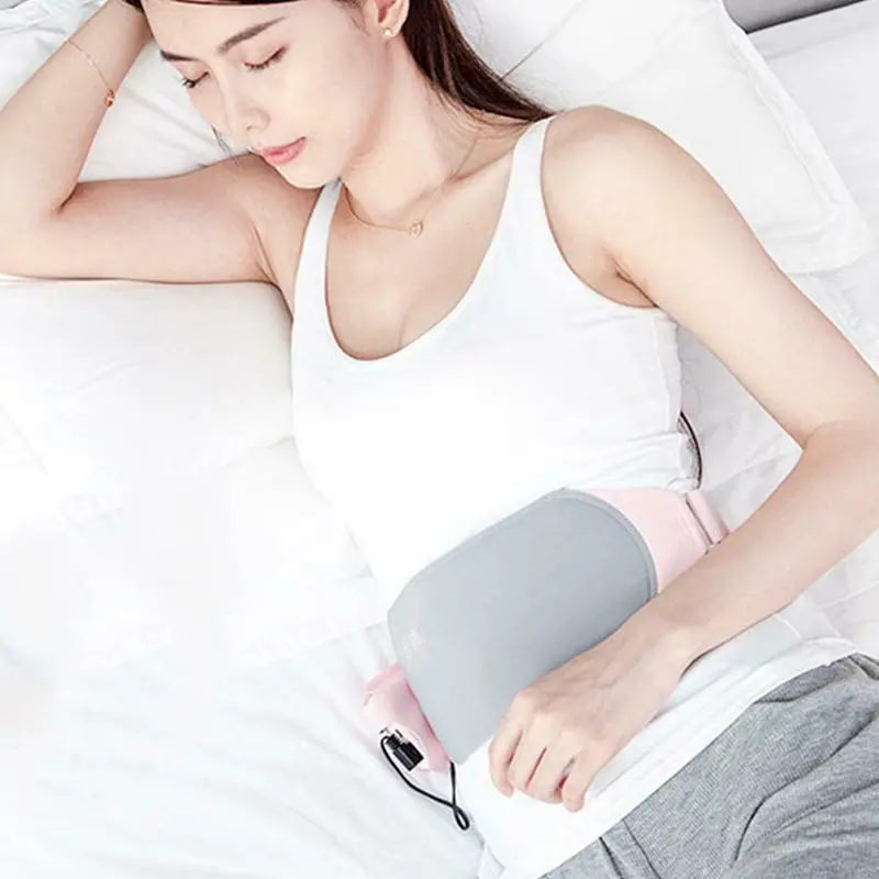 Mujer recostada en cama usando cinturón térmico eléctrico rosado y gris para aliviar dolores menstruales en el abdomen.