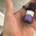 Mano sosteniendo un frasco pequeño de vidrio ámbar con tapa negra y etiqueta morada que detalla precauciones de uso para aceite esencial de aromaterapia.