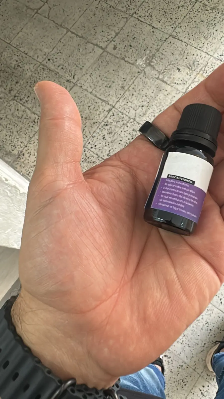 Mano sosteniendo un frasco pequeño de vidrio ámbar con tapa negra y etiqueta morada que detalla precauciones de uso para aceite esencial de aromaterapia.