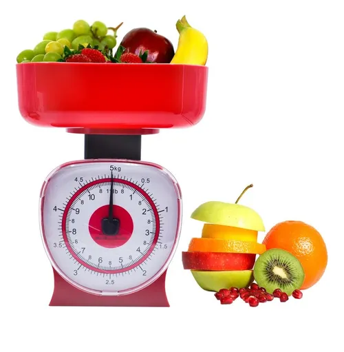 Balanza de cocina mecánica roja con bol y dial analógico de 5 kg, junto a frutas variadas sobre fondo blanco.