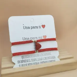 Dos manillas tejidas en hilo rojo con dije de corazón magnético, presentadas en tarjeta blanca con mensaje romántico sobre una base de madera clara.