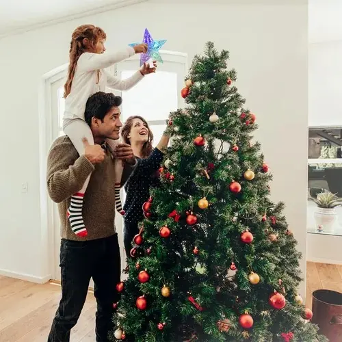 Niña sobre los hombros de su padre coloca la estrella de Navidad transparente con luces LED multicolor en la punta de un árbol decorado.