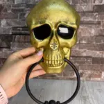 Timbre de calavera dorado y texturizado para Halloween con ojos negros profundos, altavoz nasal y aro móvil negro sujeto con la mano.