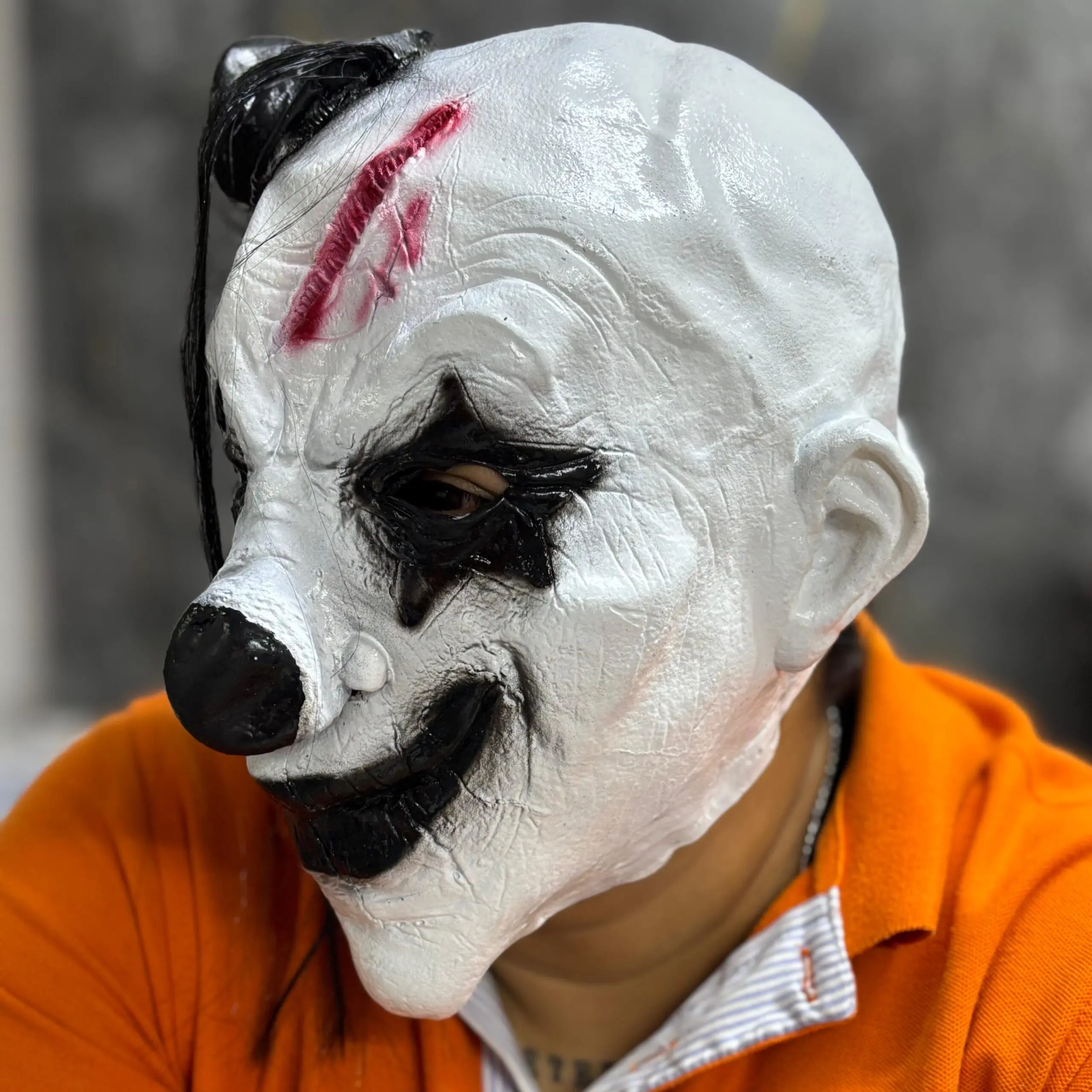 Máscara de látex de payaso aterrador en color blanco, con nariz y maquillaje de ojos negro, herida sangrienta en la frente y mechón de cabello negro.