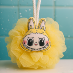 Esponja de baño amarilla de malla con rostro de Labubu, diseño de conejo sonriente y cordón blanco para colgar frente a azulejos azules.