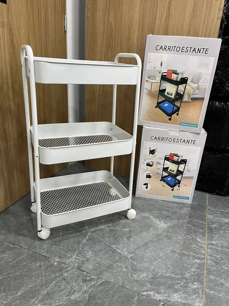 Carrito organizador blanco de tres niveles con bandejas de rejilla y ruedas, junto a sus cajas de empaque sobre piso de baldosa gris.
