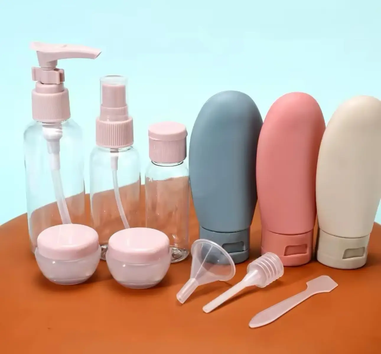 Kit de 11 piezas para viaje con tres botellas transparentes con bomba, atomizador y tapa rosa, tres frascos de silicona suaves en tonos azul, rosa y beige, dos jarras pequeñas, un embudo, una pipeta y una espátula sobre superficie marrón.