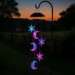 Móvil solar decorativo con estrellas y lunas LED RGB en tonos azul y violeta, colgado de un soporte metálico en un jardín al atardecer.