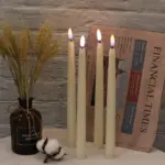 Set de cuatro velas LED cónicas de cera real color crema con luz cálida, dispuestas frente a un periódico, un florero con espigas y una flor de algodón.