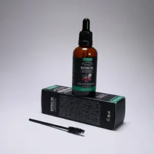 Frasco ámbar con gotero de aceite de batana sobre su caja negra y verde, acompañado de un cepillo aplicador negro.