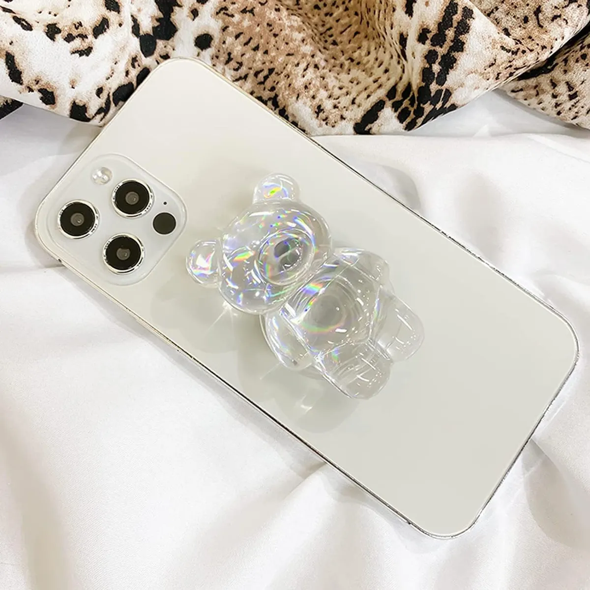 Pop socket de acrílico transparente con forma de osito iridiscente en un celular blanco sobre tela blanca y estampado de leopardo.