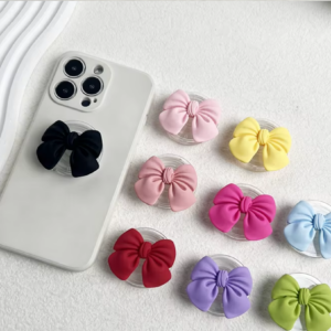 Pop sockets con moños de tela de colores y base de acrílico. Un modelo negro en celular blanco y varios tonos rosa, amarillo, azul y verde.