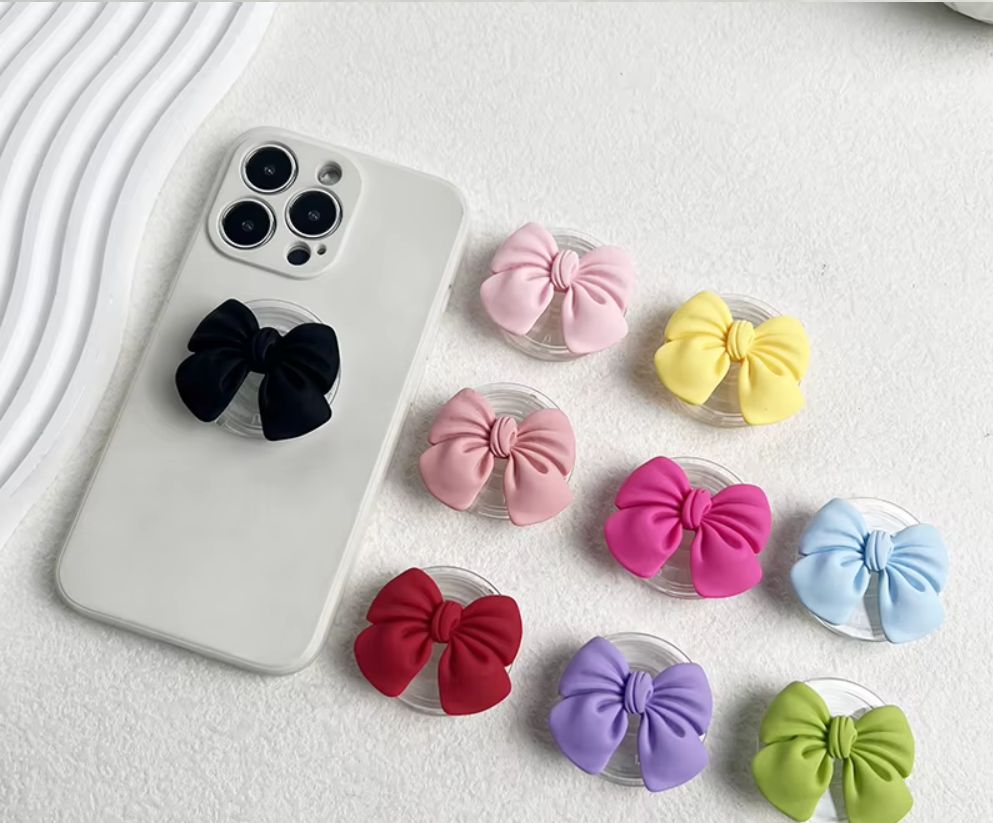 Pop sockets con moños de tela de colores y base de acrílico. Un modelo negro en celular blanco y varios tonos rosa, amarillo, azul y verde.