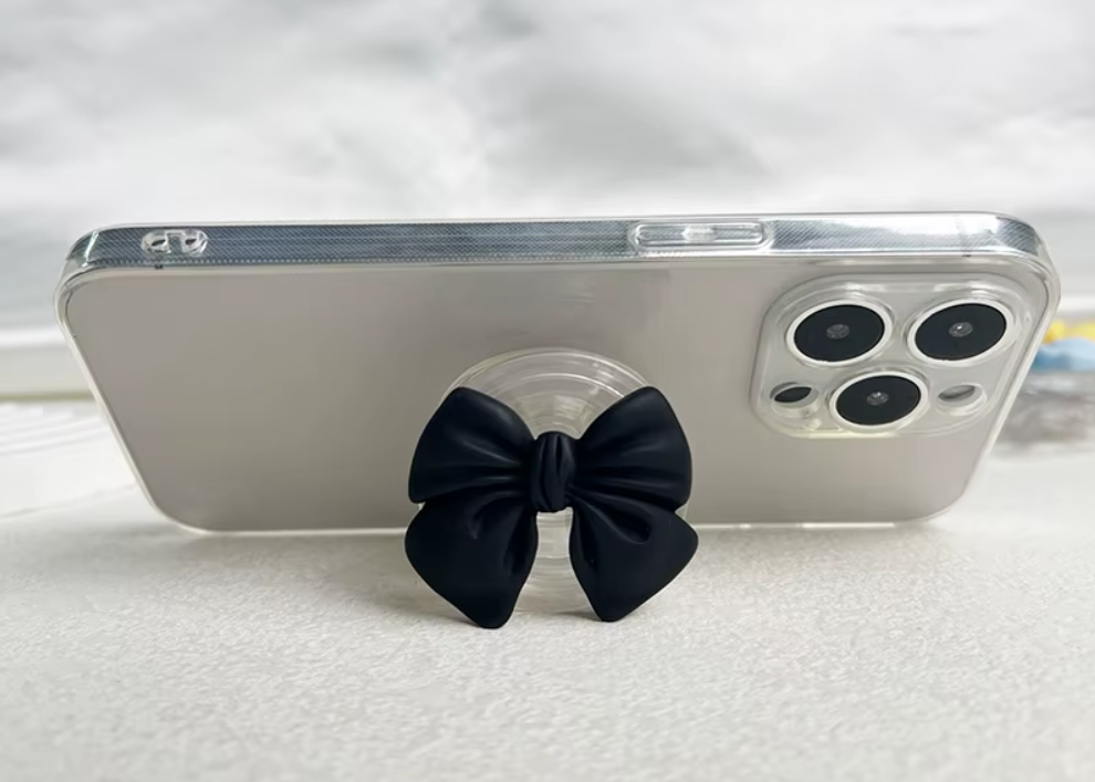 Pop socket de tela negra con diseño de moño para celular, instalado en un teléfono con estuche transparente sobre una superficie blanca.