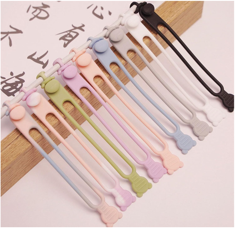 Sujetadores multipropósito de silicona flexible en diversos colores pastel y neutros, dispuestos en diagonal sobre una superficie de madera.